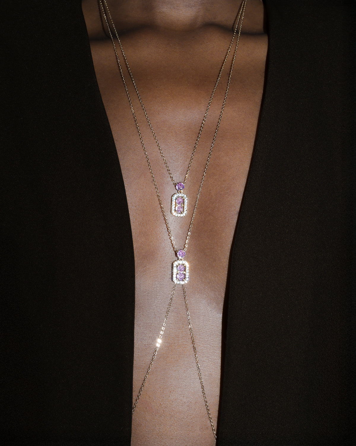 A close-up of a model's neck and upper chest wearing a layered gold necklace and body chainwith two small rectangular purple gemstone pendants surrounded by clear stones, set against a dark background.