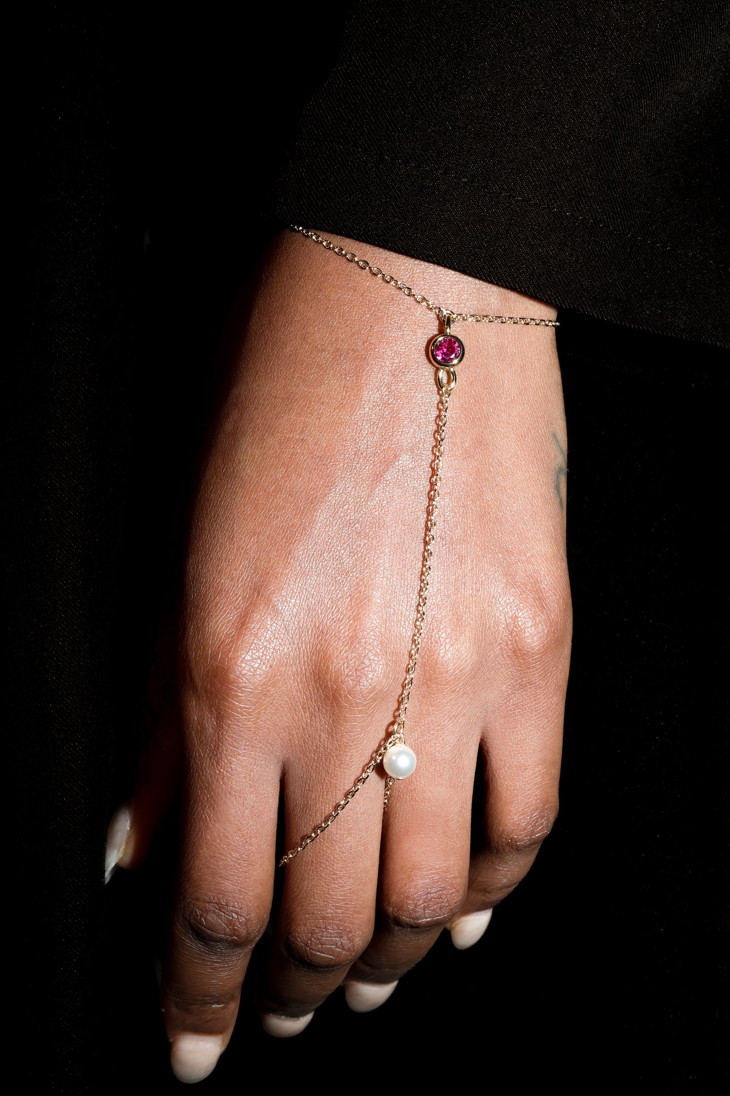 Hand wearing a delicate gold bracelet with a red gemstone on a black background