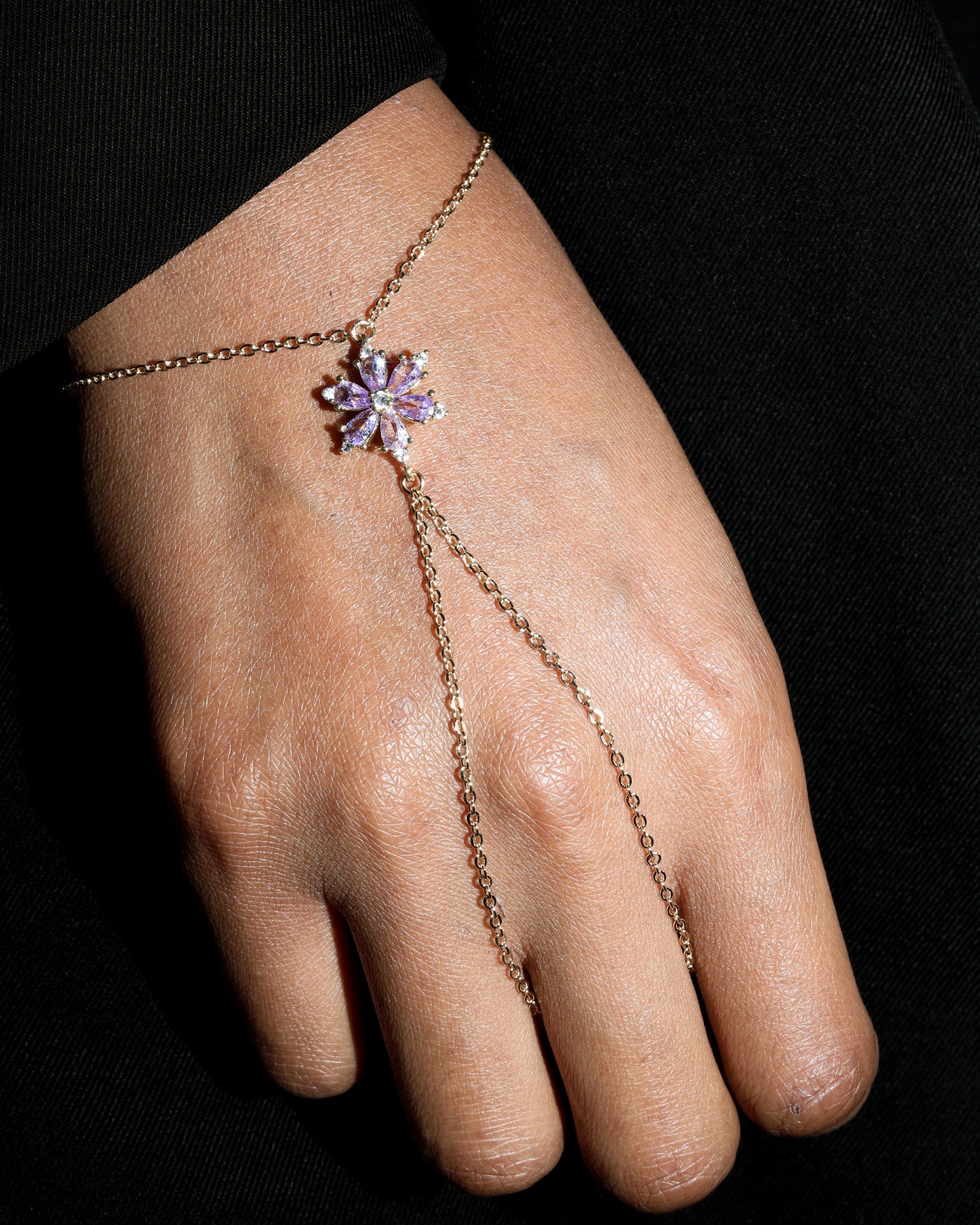 Hand wearing a delicate gold hand chain with a purple floral crystal charm on a black background