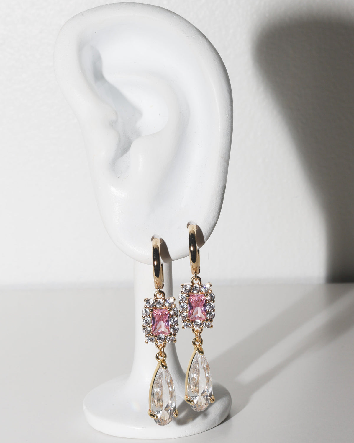 A pair of gold drop earrings with pink gemstones and clear teardrop crystals, displayed on a white ear-shaped jewelry stand against a white background.