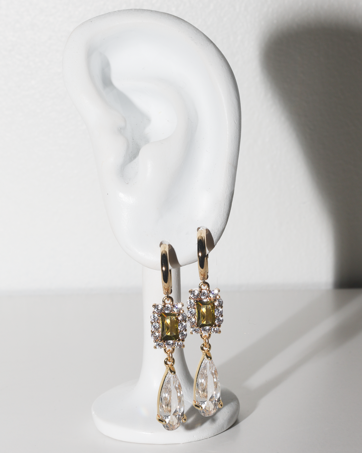 A pair of gold drop earrings with rectangular olive-green stones and clear teardrop crystals, displayed on a white ear-shaped jewelry stand against a white background.