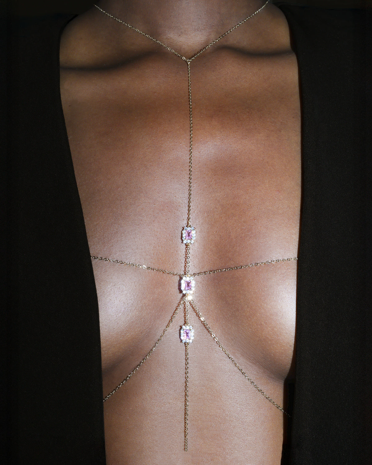 A close-up of a model's bare chest wearing a delicate gold chain body chain with three rectangular pink gemstones, set against a black background.