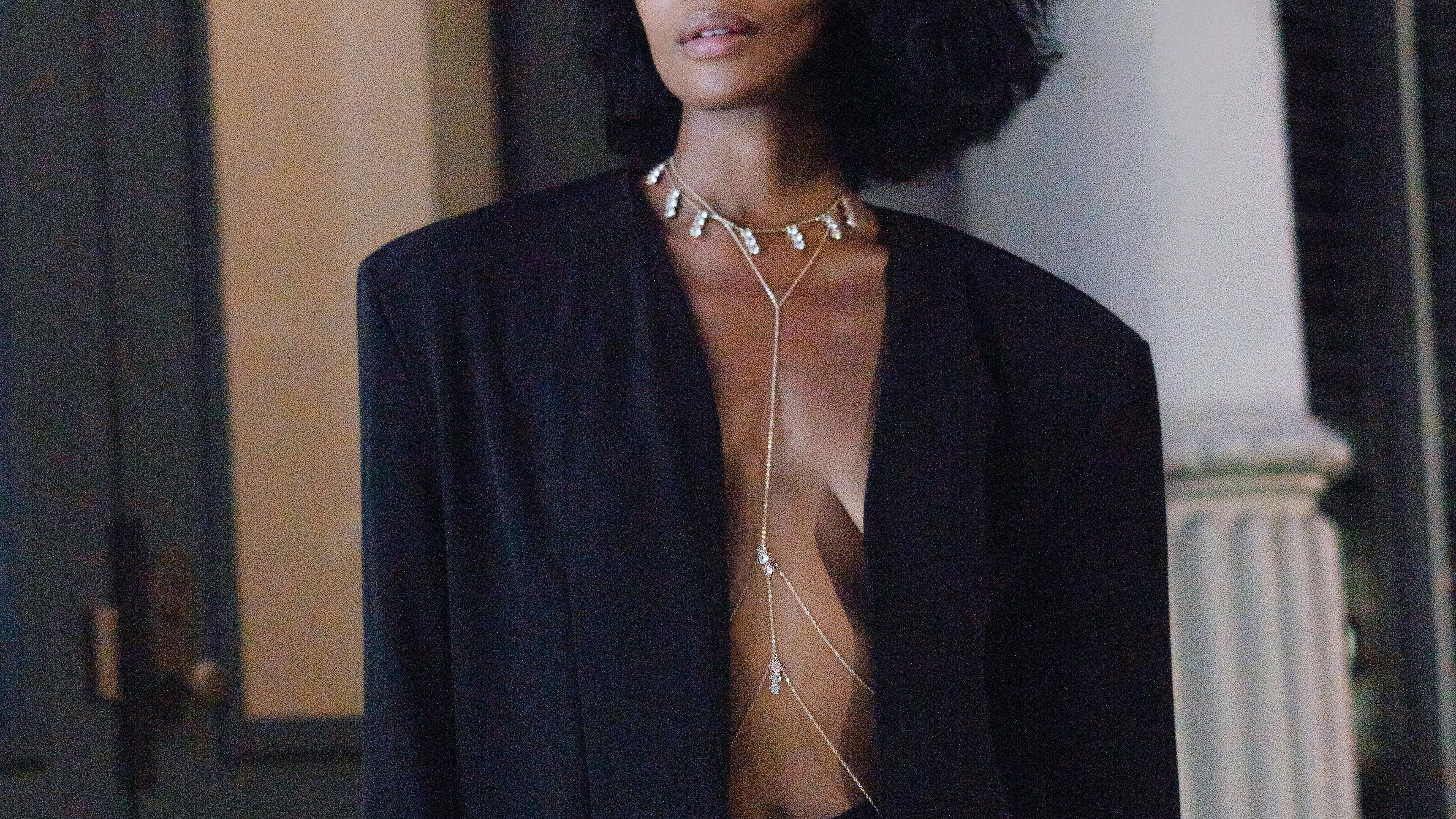 Woman wearing a black blazer over a sheer top with a necklace and body chain , standing indoors.