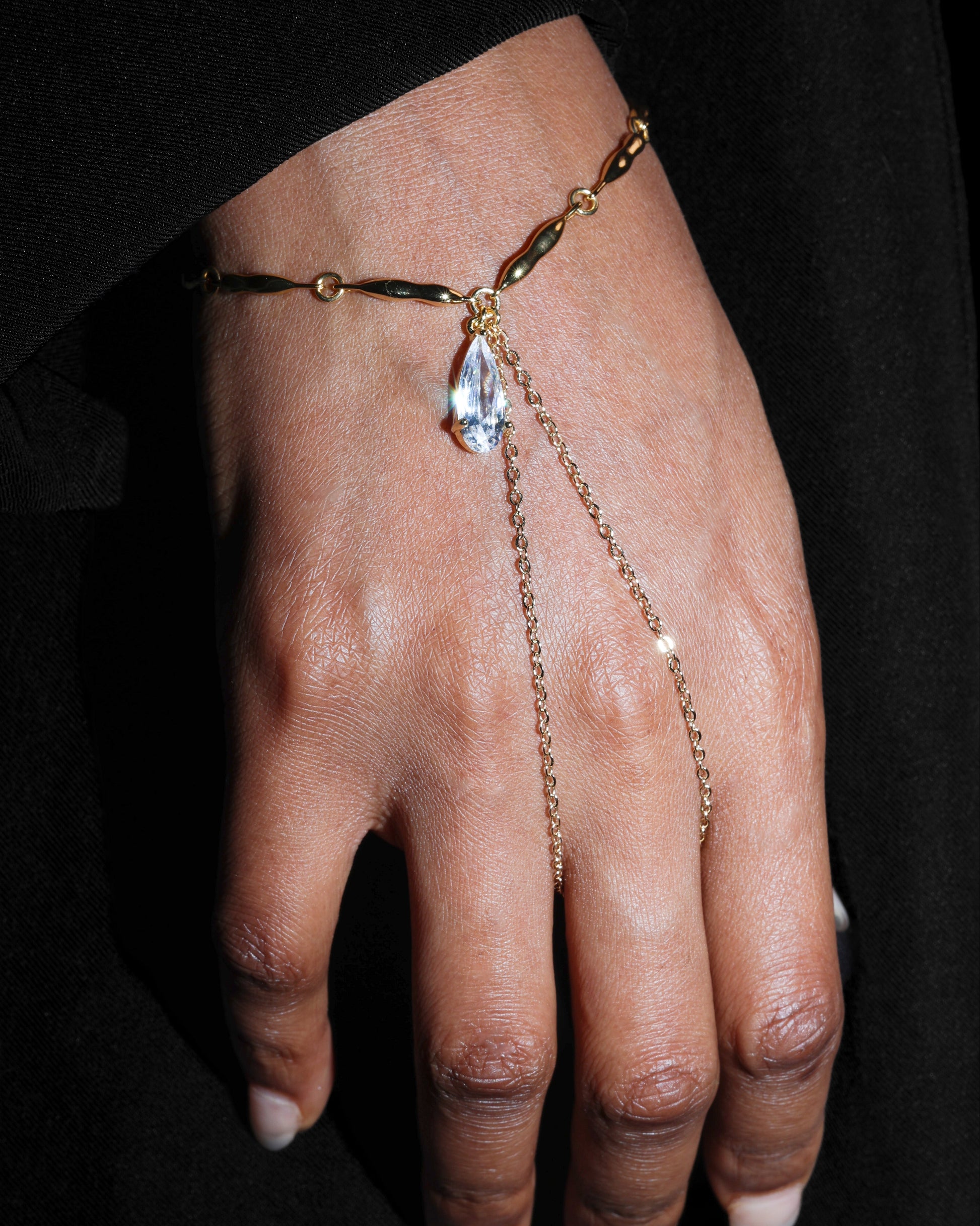 Hand wearing a delicate gold bracelet with a clear gemstone on a black background