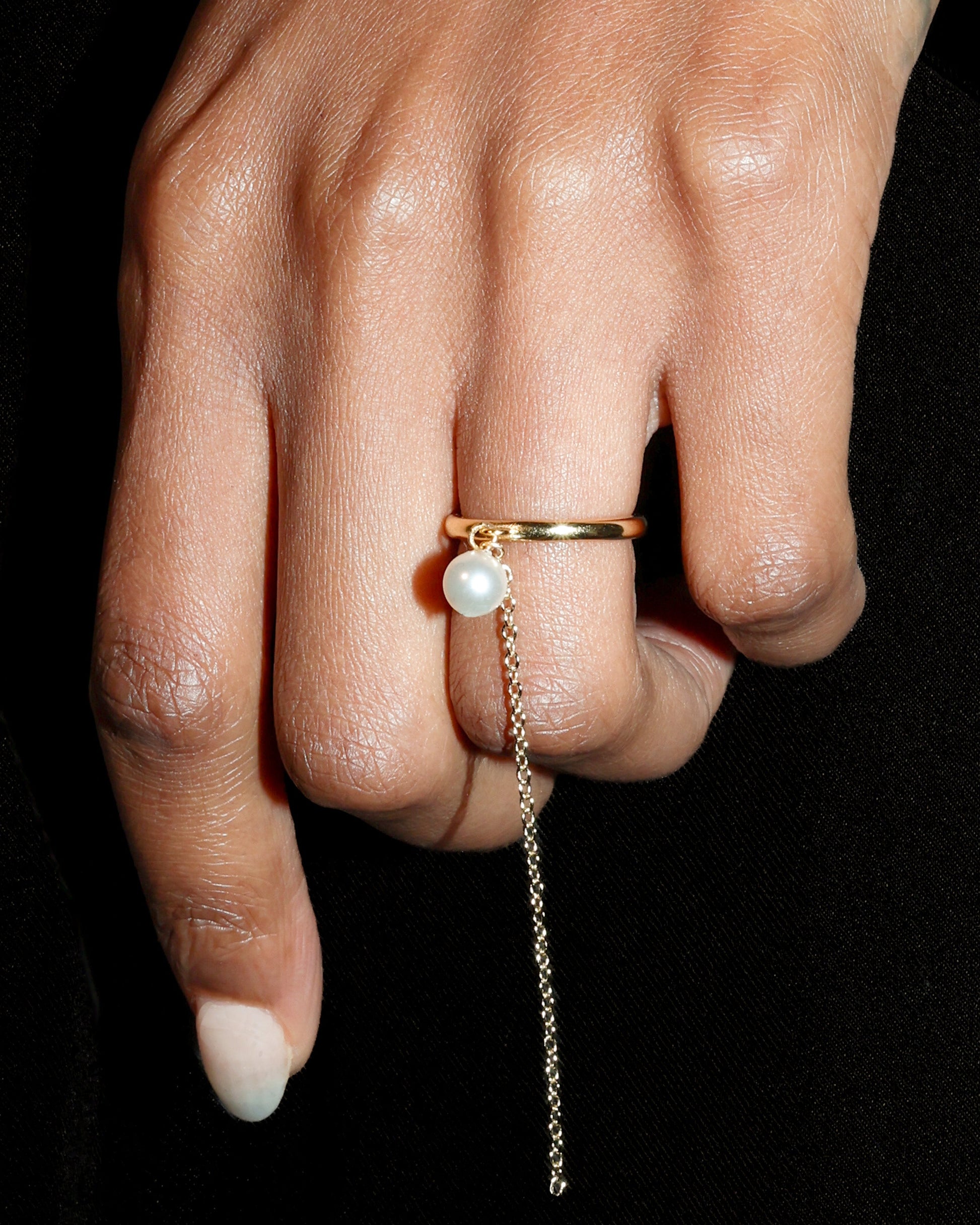 Hand wearing a gold ring with a pearl and a dangling chain on a black background