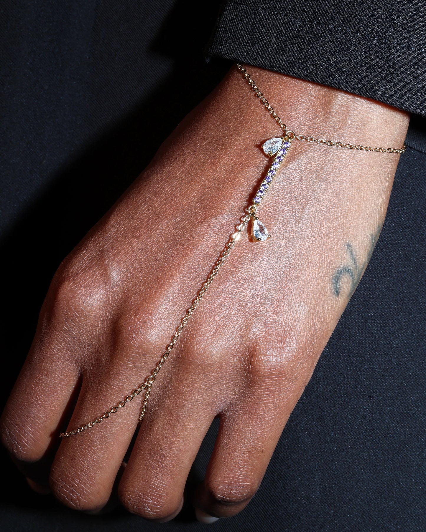Hand wearing a delicate gold hand harness bracelet with small purple stones against a dark background