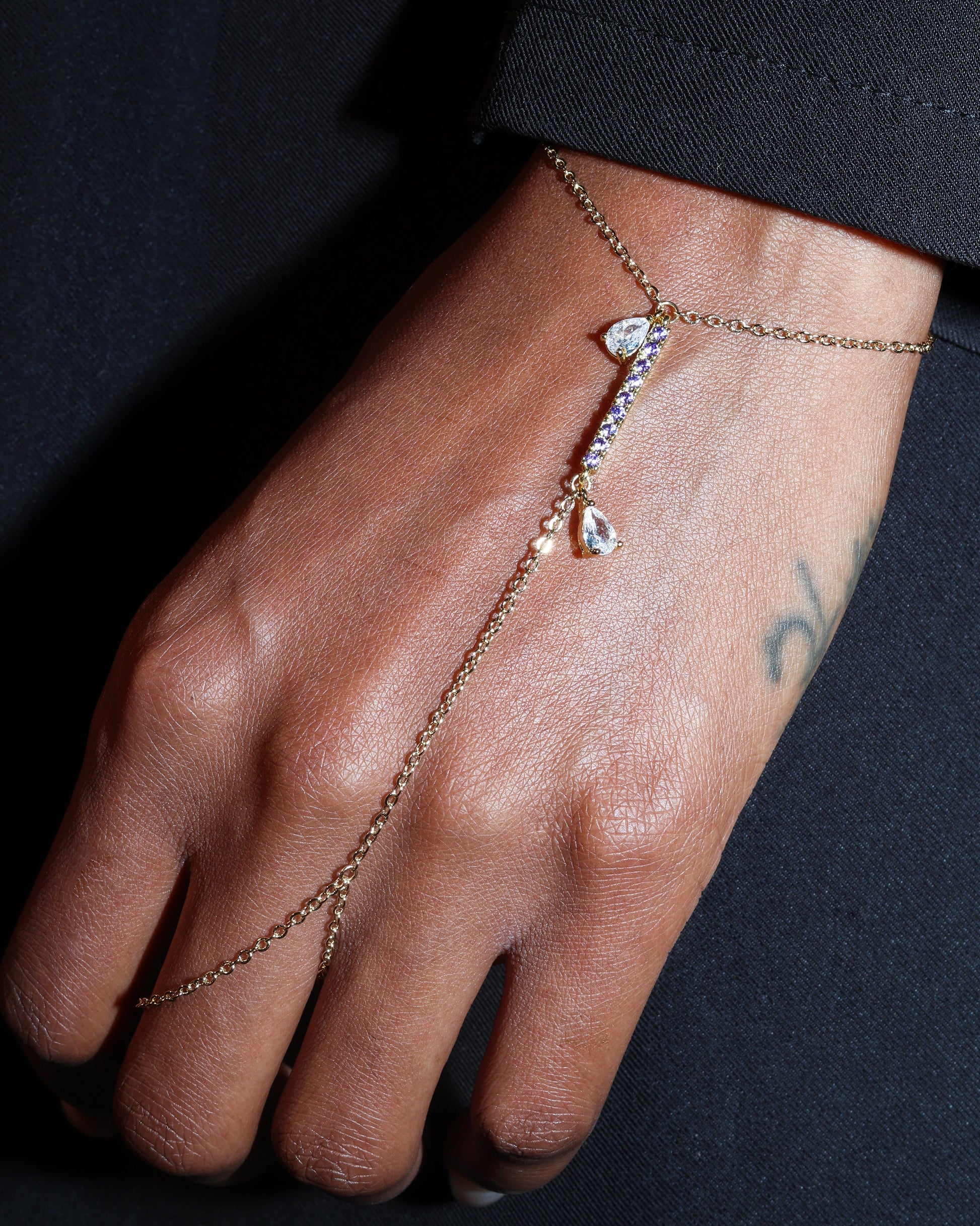 Hand wearing a delicate gold hand harness bracelet with small purple stones against a dark background