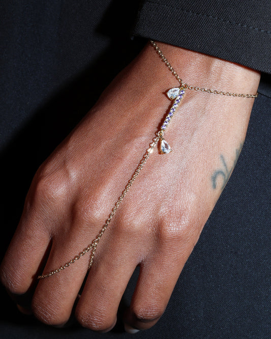 Hand wearing a delicate gold hand harness bracelet with small purple stones against a dark background