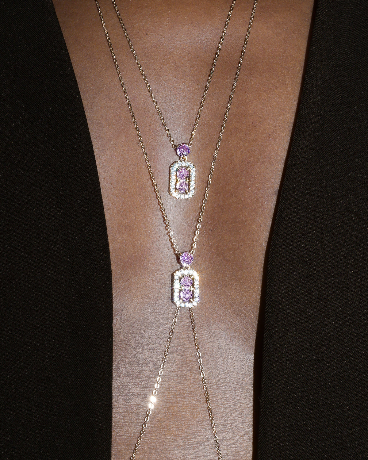 A close-up of a model's neck and upper chest wearing a layered gold necklace and body chainwith two small rectangular purple gemstone pendants surrounded by clear stones, set against a dark background.
