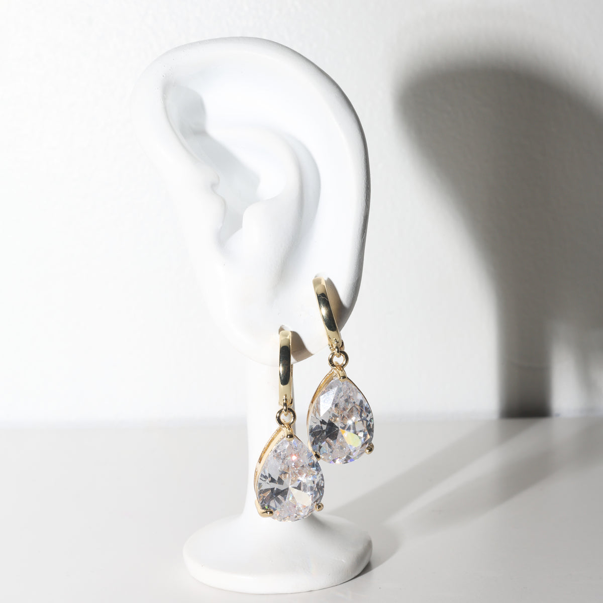 A pair of gold hoop earrings with large clear teardrop-shaped rhinestones, displayed on a white ear-shaped stand against a white background.