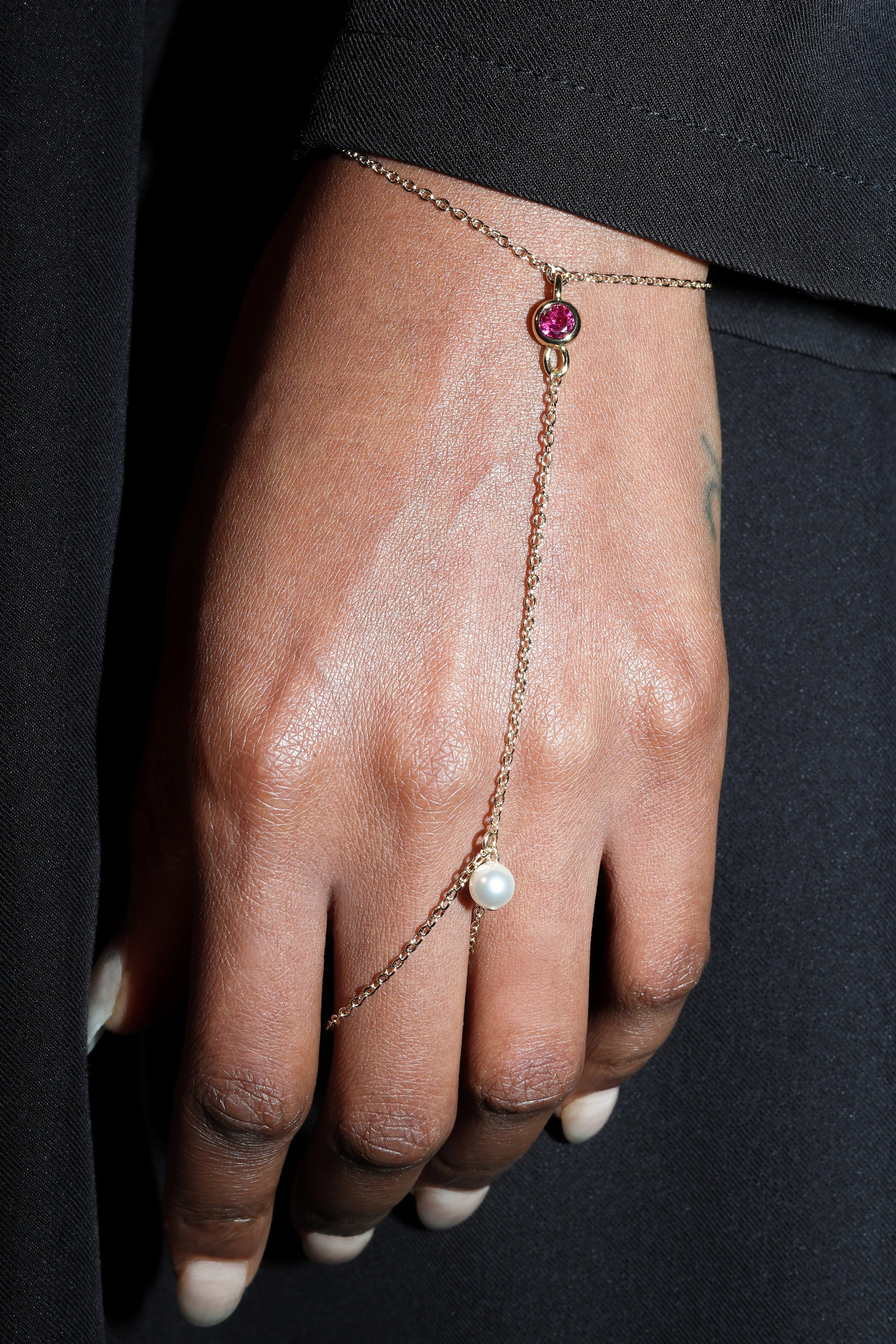 Hand wearing a delicate gold hand harness with a pink gemstone and pearl on a black background