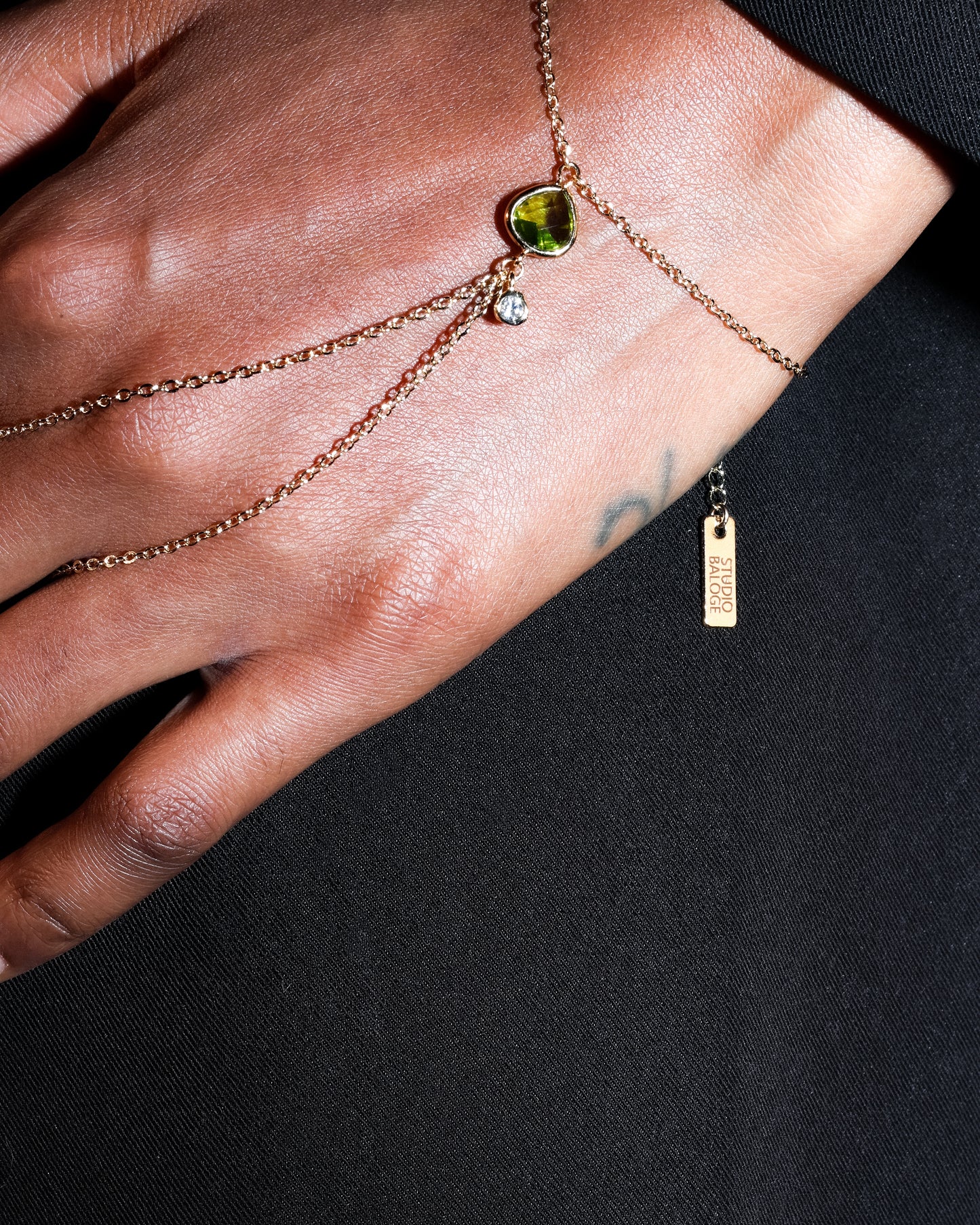 Close-up of a hand wearing the Point Hand Harness in 14k gold-filled chain with peridot gemstone drop on a black background 