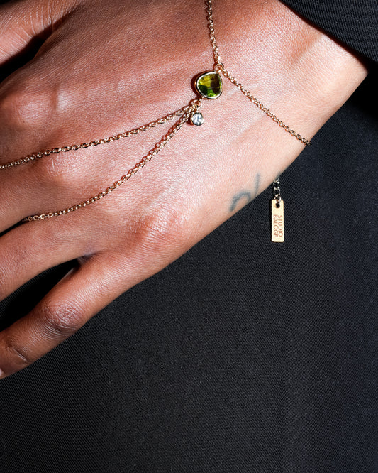 Close-up of a hand wearing the Point Hand Harness in 14k gold-filled chain with peridot gemstone drop on a black background 