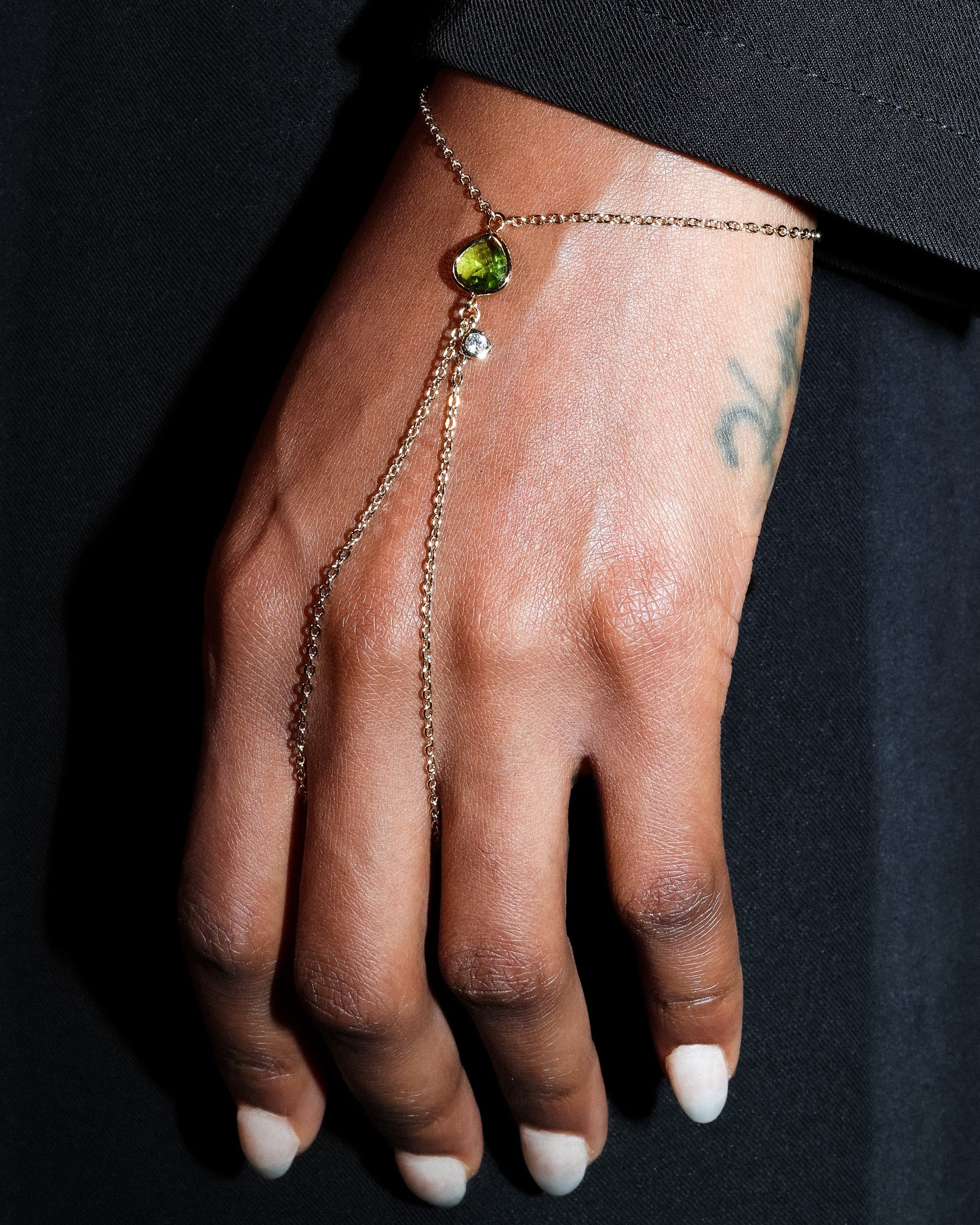 Hand wearing a gold hand harness bracelet with a green gemstone on a black background