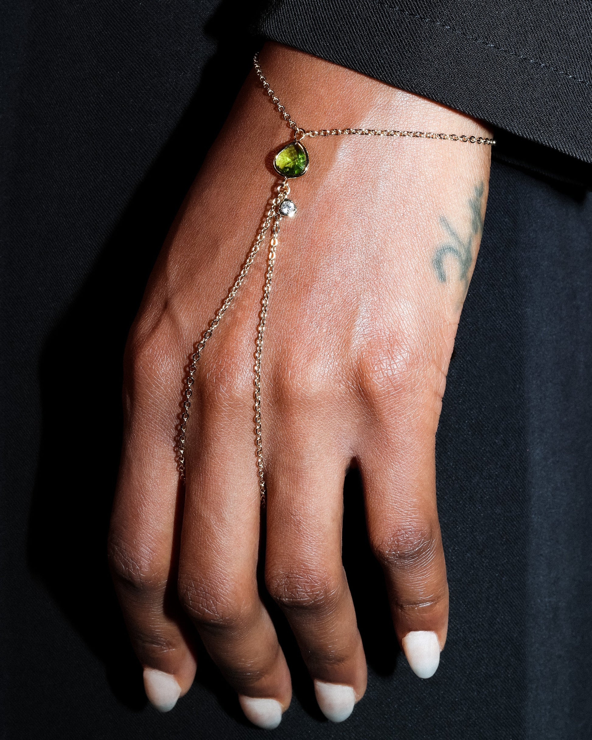 Hand wearing a gold hand harness bracelet with a green gemstone on a black background