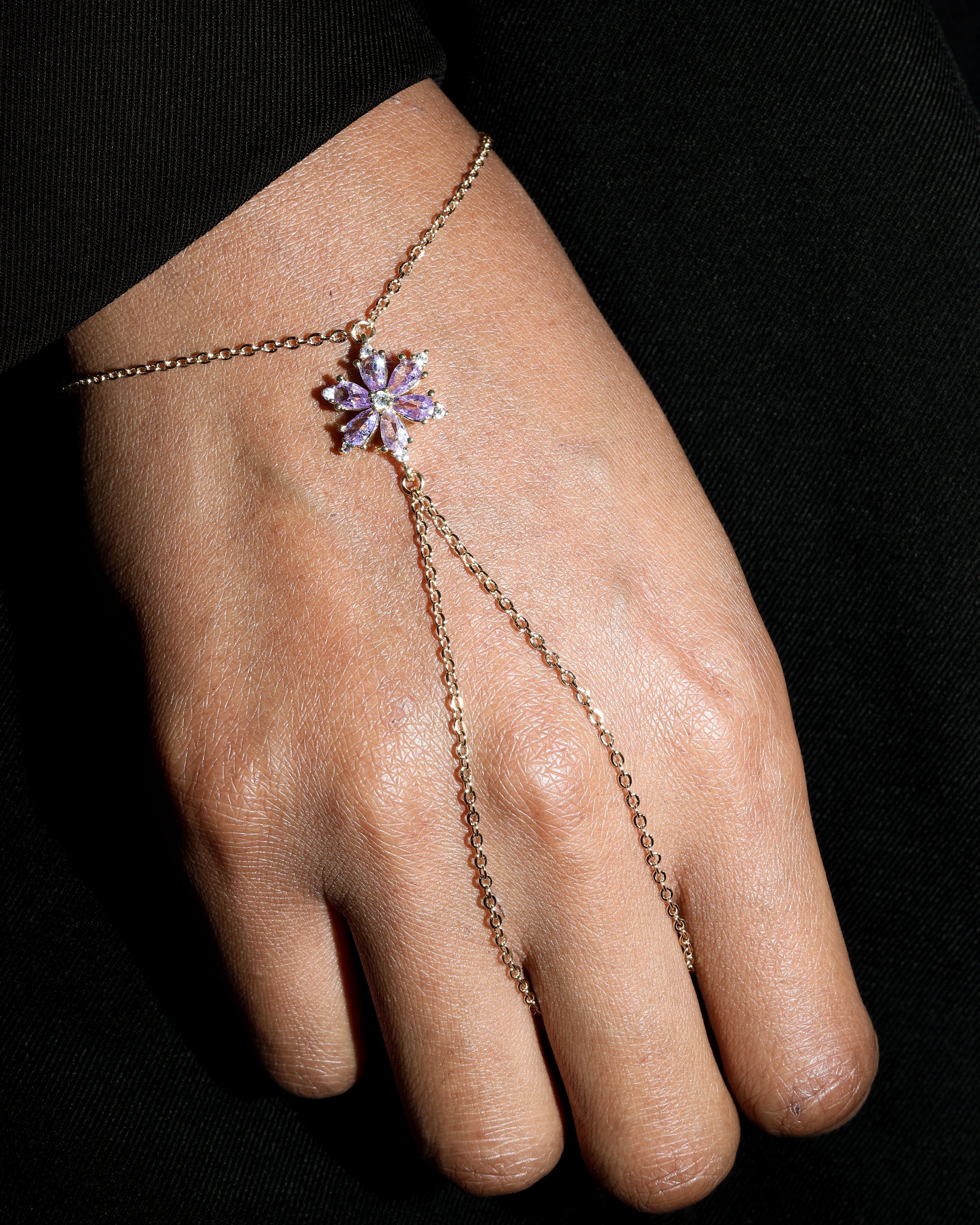 Hand wearing a delicate gold hand chain with a purple floral crystal charm on a black background