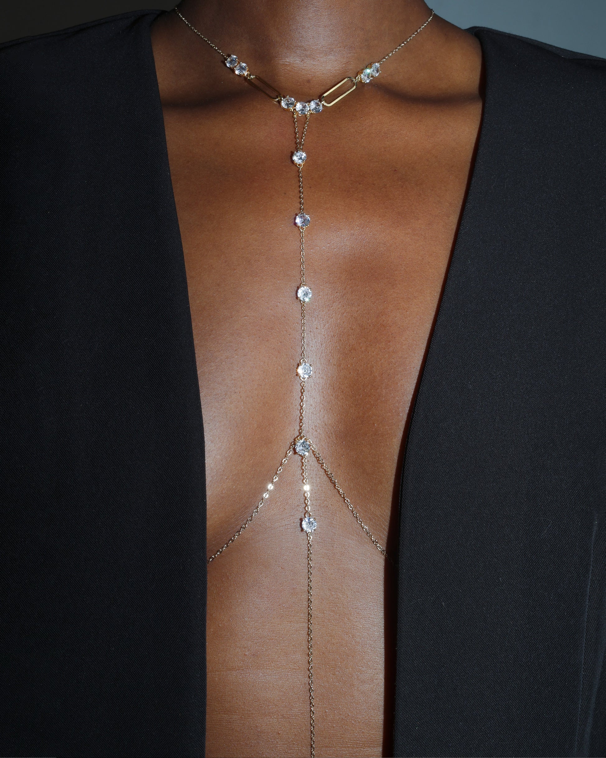 A close-up of a model wearing a delicate gold-toned chain choker necklace with clear rhinestones that extend down the chest like a harness, styled with a black open-front blazer.