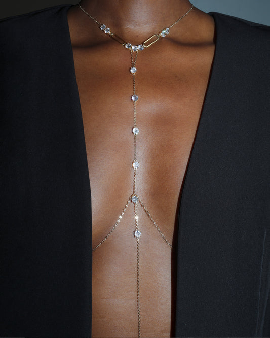 A close-up of a model wearing a delicate gold-toned chain choker necklace with clear rhinestones that extend down the chest like a harness, styled with a black open-front blazer.