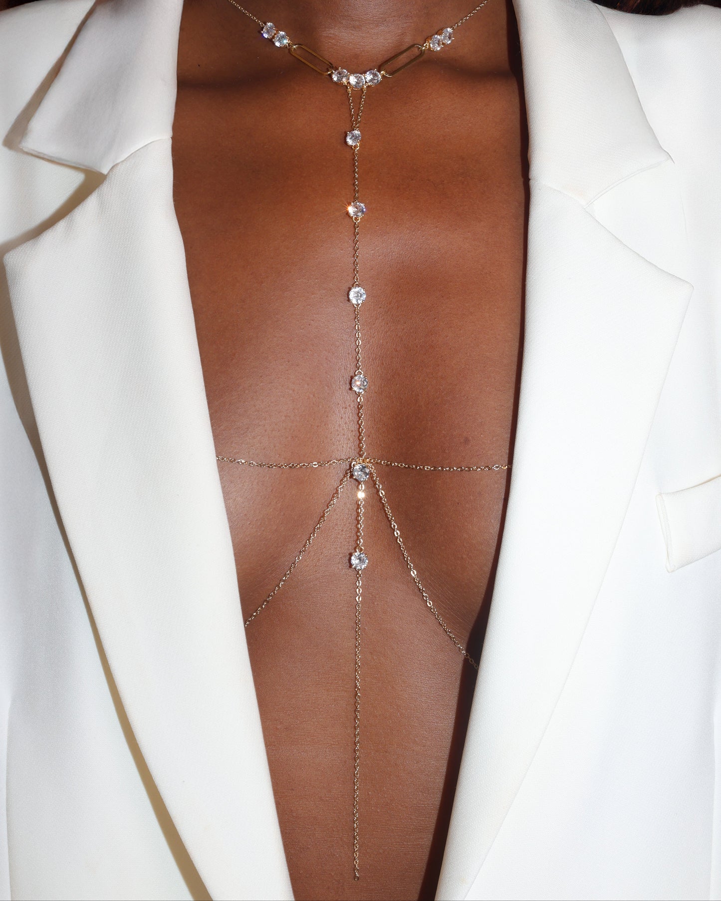 A close-up of a model wearing a delicate gold body chain with clear rhinestones, styled with a white blazer.