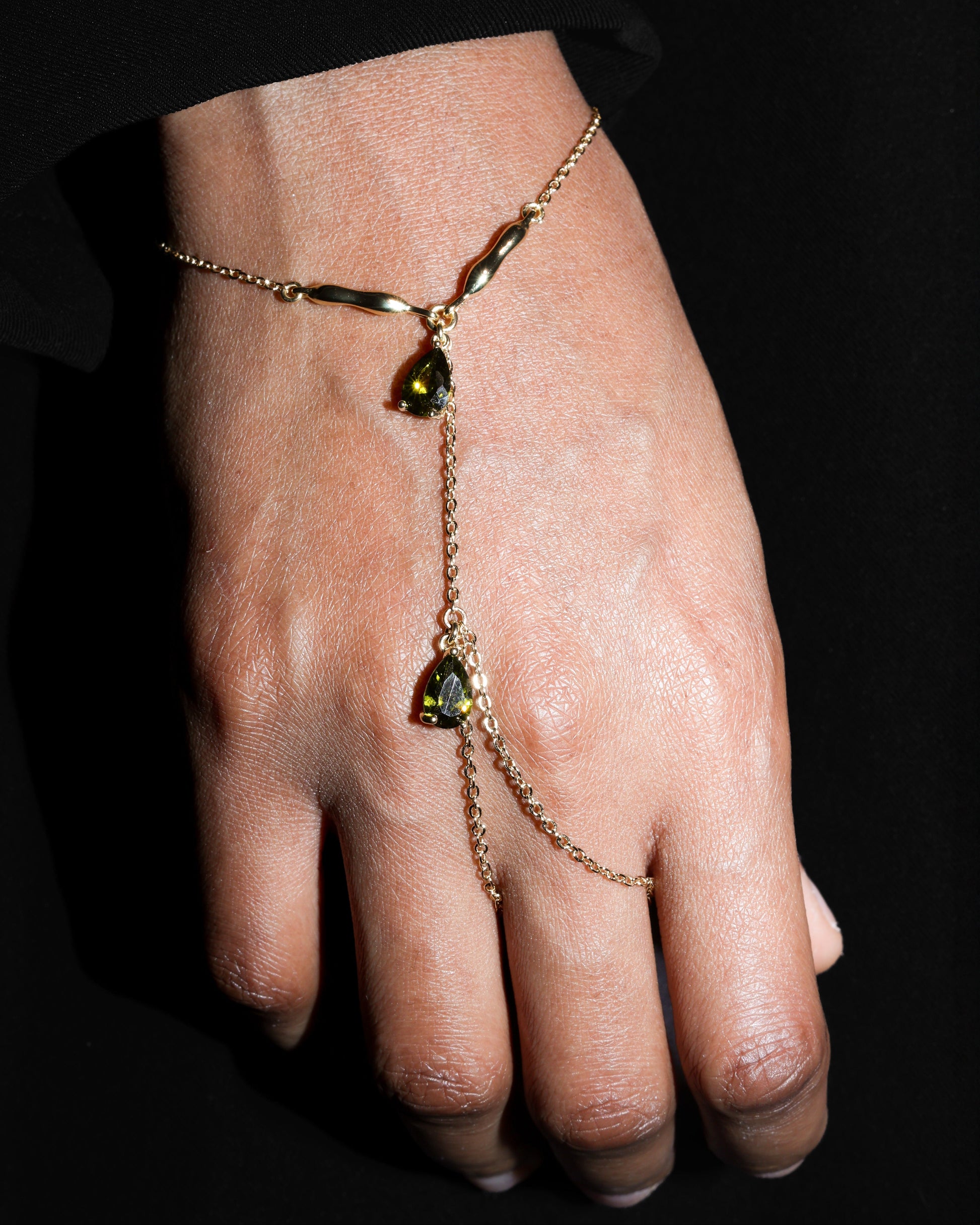 Hand wearing a delicate gold bracelet with  olive green gemstones against a dark background