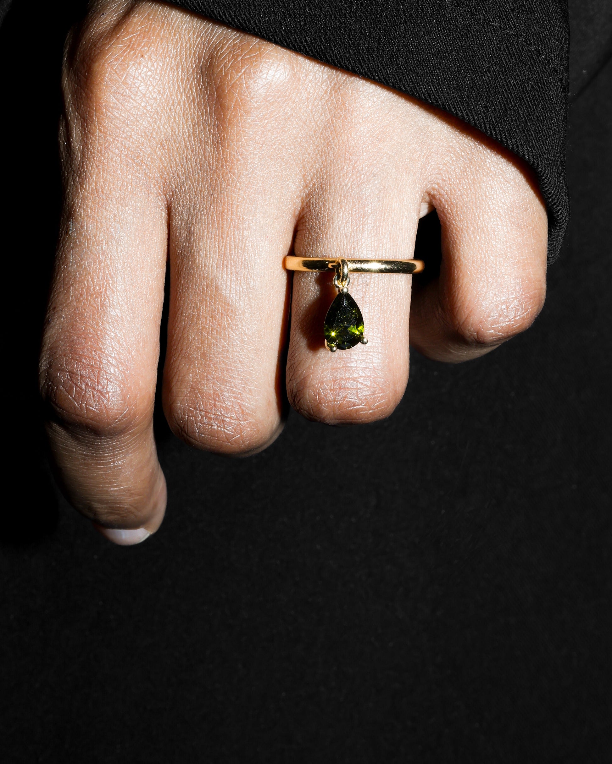 Hand wearing a gold ring with a green gemstone against a black background