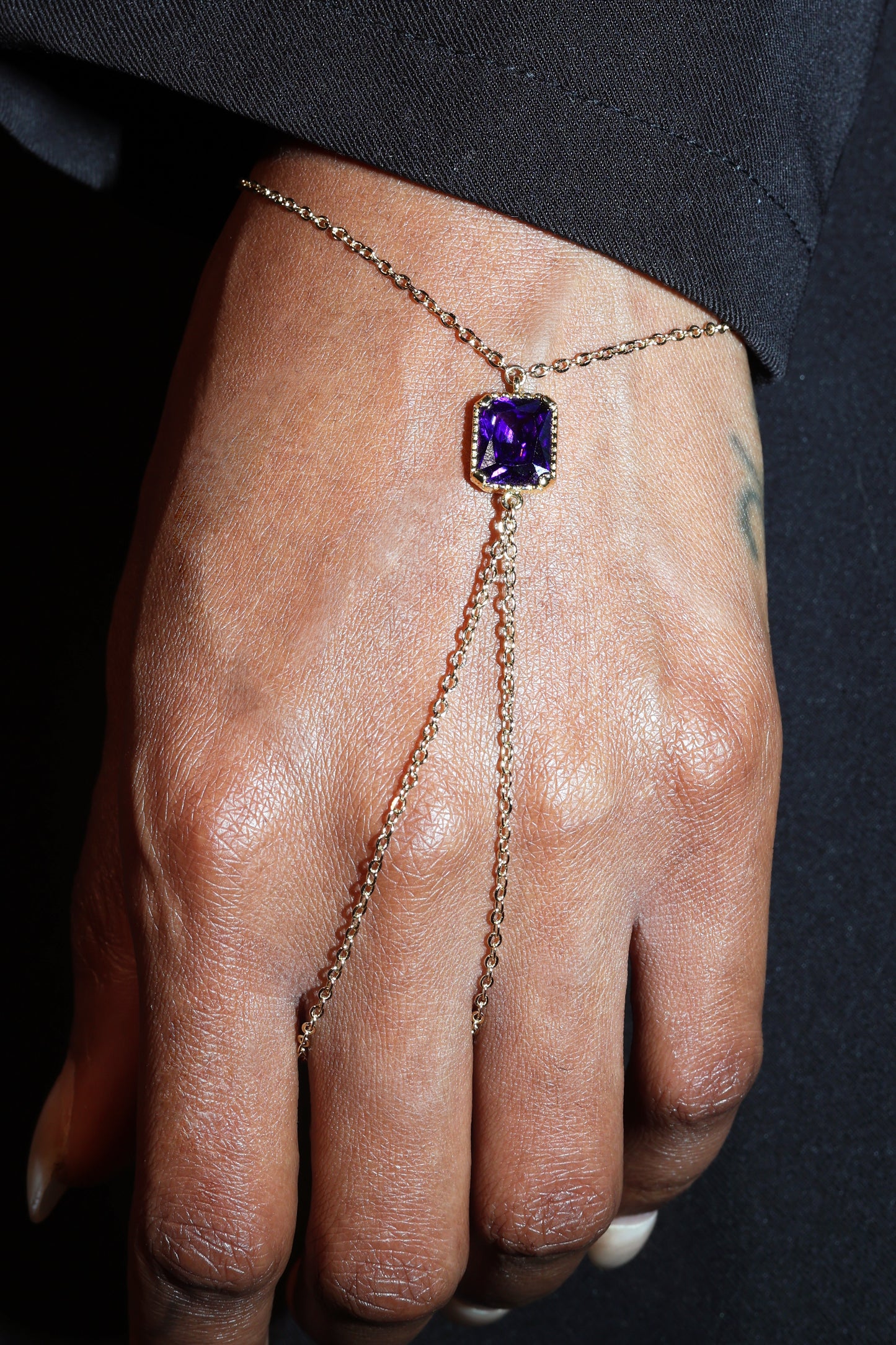 Hand wearing a gold chain bracelet with a purple gemstone on a dark background