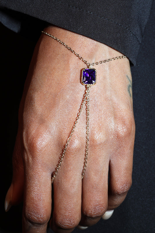 Hand wearing a gold chain bracelet with a purple gemstone on a dark background